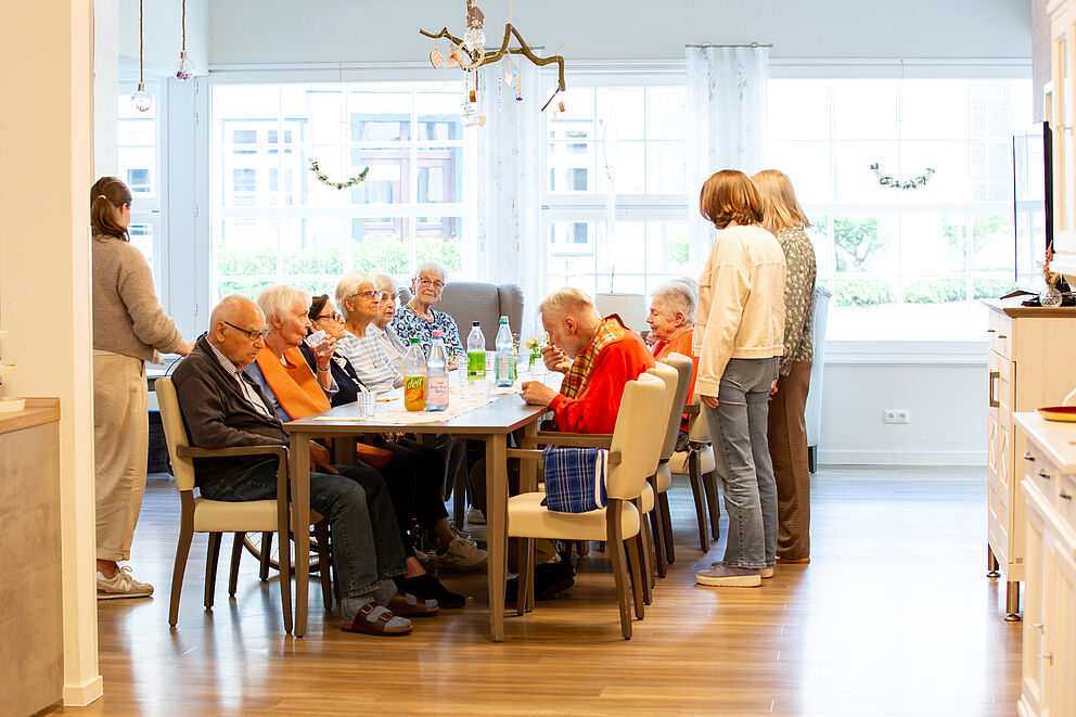 Mehrere ältere Menschen sitzen an einem Tisch in einem hellen Raum, zwei jüngere Personen stehen daneben.
