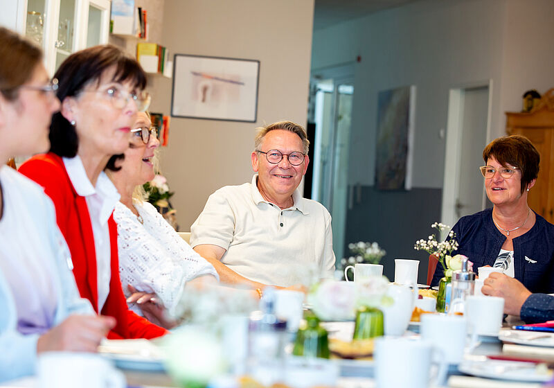 Gruppe von Menschen sitzt an einem Tisch mit Tassen und Pflanzen und unterhält sich.