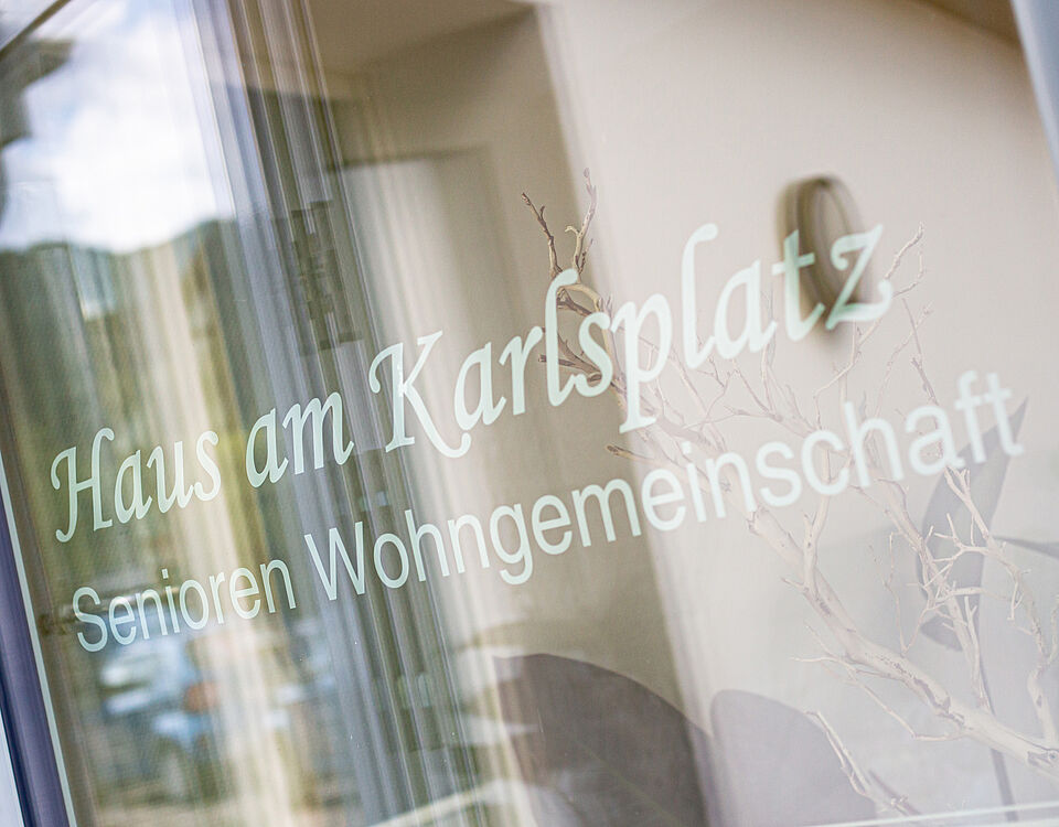 Fenster mit klar lesbarer Aufschrift "Haus am Karlsplatz - Senioren Wohngemeinschaft" und Spiegelung der Umgebung.