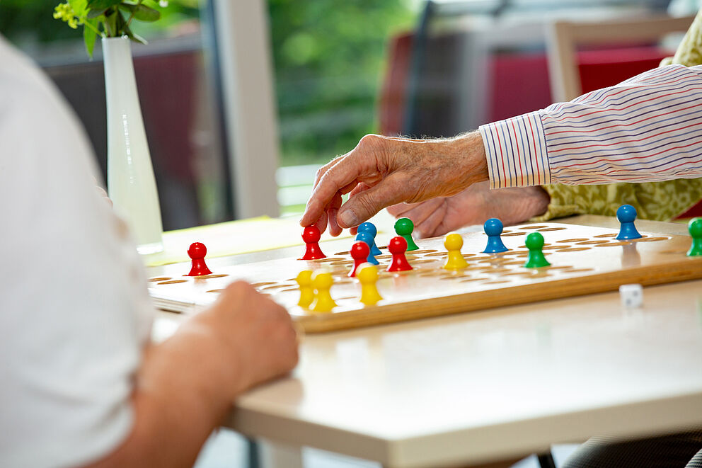 Zwei Personen spielen mit bunten Spielfiguren auf einem Holzbrett an einem weißen Tisch.