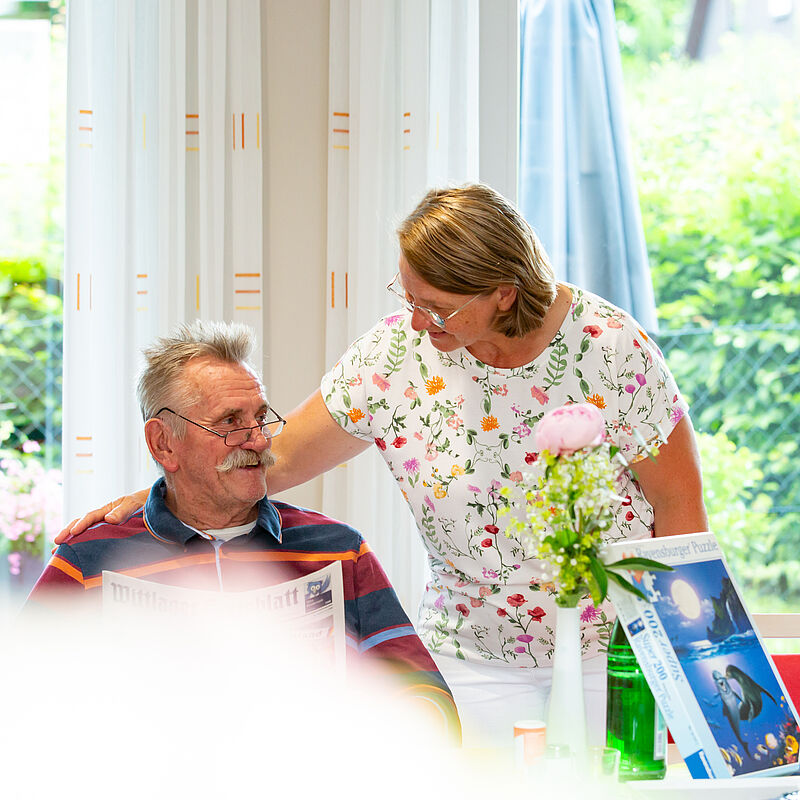 Älterer Mann mit Brille sitzt und liest Zeitung während eine Frau mit kurz geschnittenem Haar und geblümtem T-Shirt sich zu ihm beugt und ihn anlächelt.