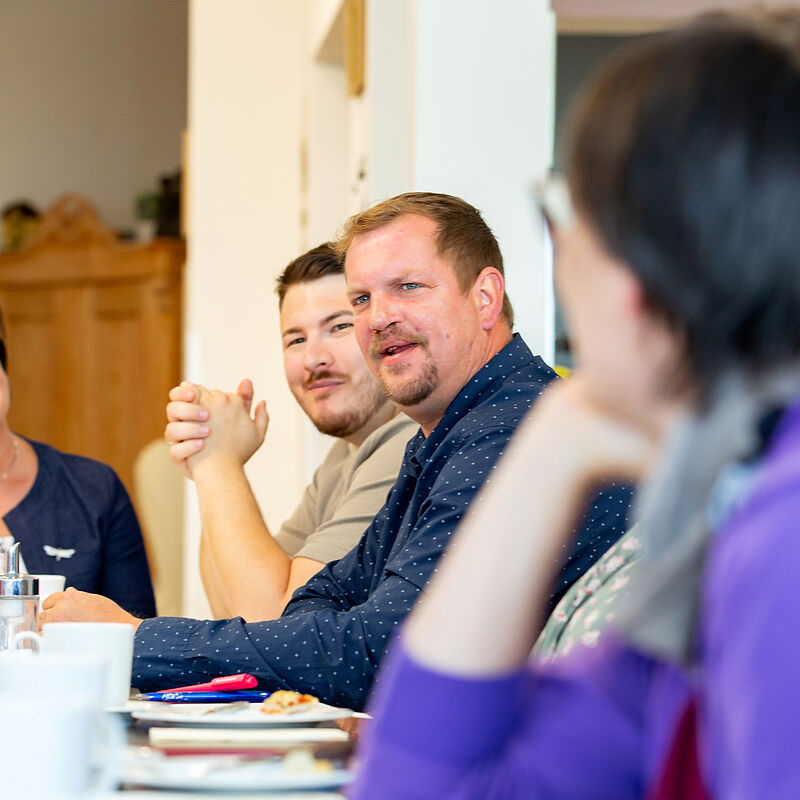Gruppe von Menschen sitzt an einem Tisch und unterhält sich, im Vordergrund eine sitzende, unscharfe Person mit lila Pullover.