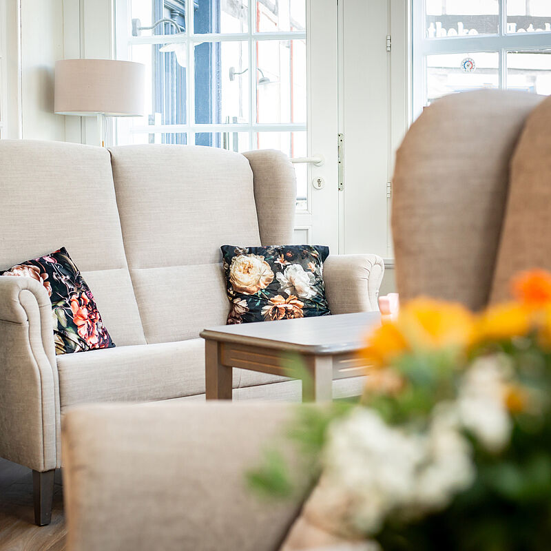 Beiges Sofa mit floral gemusterten Kissen vor einem Fenster und einem kleinen Holztisch.