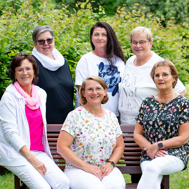 Sechs Frauen unterschiedlichen Alters sitzen und stehen in einem Garten vor grünen Büschen, drei sitzen auf einer Bank, drei stehen dahinter, alle lächeln.