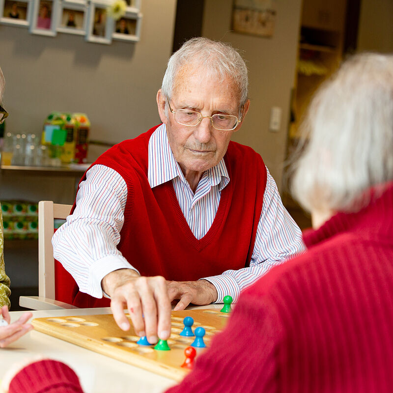 Drei ältere Menschen sitzen an einem Tisch und spielen ein Brettspiel mit bunten Spielfiguren.