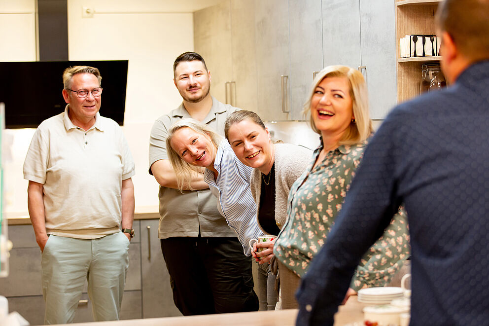 Fünf Personen stehen in einer Küche und lachen eine sechste Person an, die mit dem Rücken zur Kamera steht.