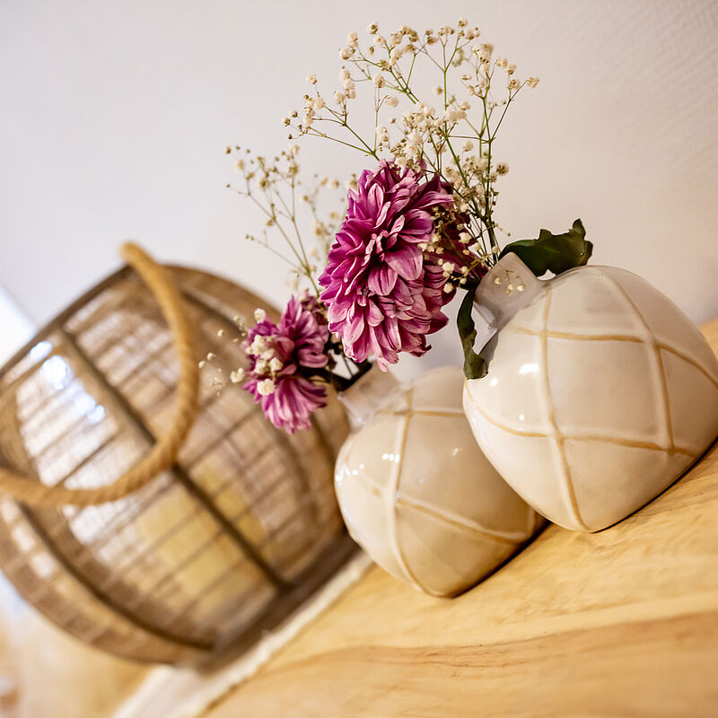 Zwei weiße Vasen mit Gittermuster auf einem Holztisch, darin rosa Blumen und kleine weiße Blüten, im Hintergrund ein geflochtener Korb.