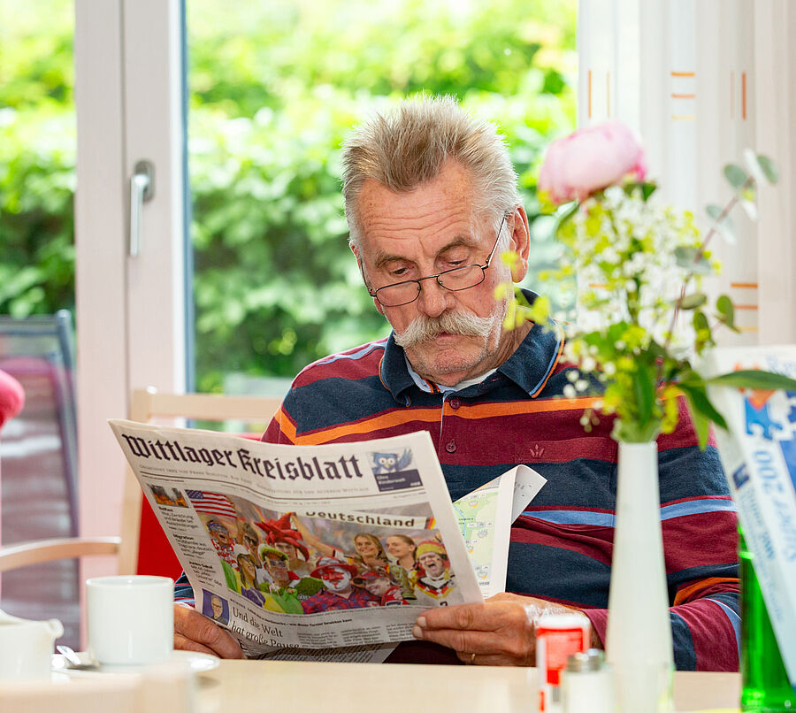 Älterer Mann mit Brille und gestreiftem Pullover liest Zeitung in einem hellen Raum mit Fenster und Pflanzen.