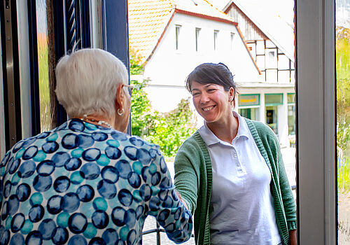 Junge Frau mit grünem Cardigan und weißem Poloshirt begrüßt ältere Frau mit weißem Haar und gemustertem Oberteil an einer offenen Haustür.