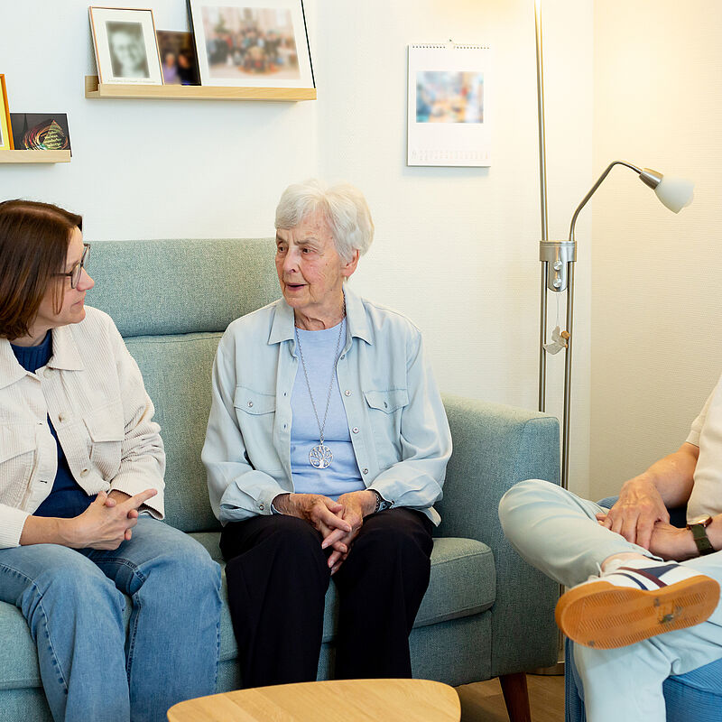 Drei Personen unterschiedlichen Alters sitzen in einem Wohnzimmer auf einem Sofa und Sessel und unterhalten sich.
