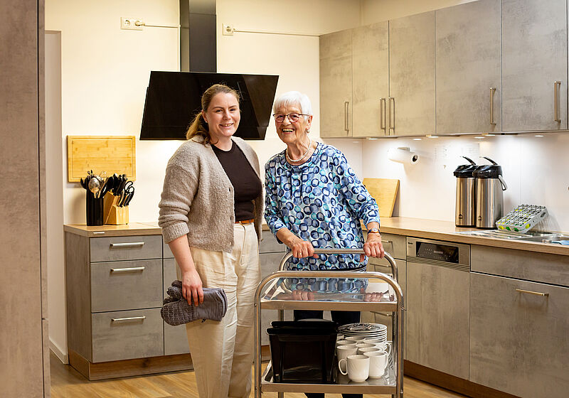 Zwei Frauen in einer modernen Küche, eine ältere mit Rollator und eine jüngere mit grauer Strickjacke, stehen lächelnd neben einem Servierwagen mit Tassen.