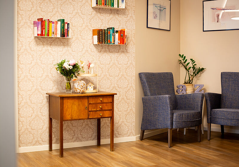 Holzkommode mit Blumenstrauß vor gemustertem Tapetenhintergrund neben zwei blauen Sesseln mit Pflanzen und Büchern an der Wand.
