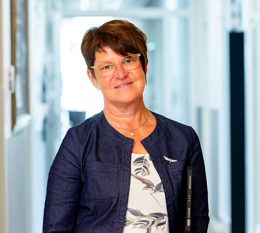 Frau mit kurzer brauner Frisur, Brille und dunkelblauem Blazer steht in einem hellen Flur und lächelt in die Kamera.